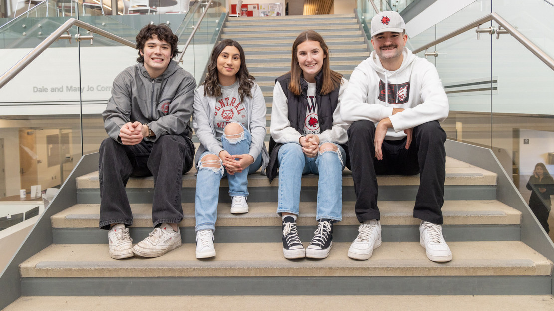Four students smile as they take a seat on the stairs inside of Samuelson Hall at Central Washinhhon University and pose for a picture.
