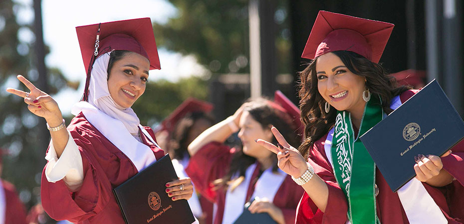 Two smiling students in CWU's crimson caps and gowns holding their diplomas.