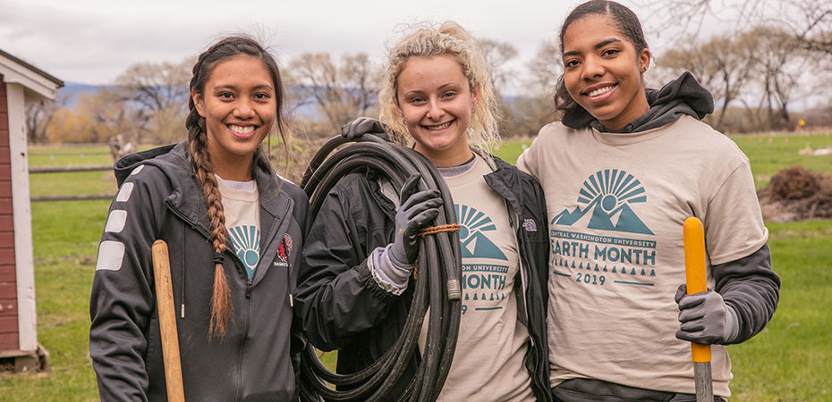 Three female students smiling wearing matching Earth Month t-shirts. 