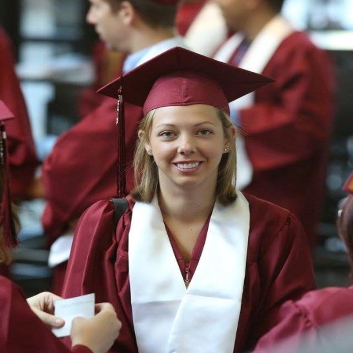Graduate in red gown smiling for the camera. She is surrounded by other graduates in the background