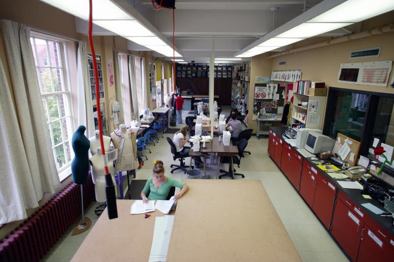 An overhead image of the costume shop. An instructor in a green shirt is sitting at a large desk and students are working at sewing machines behind her.