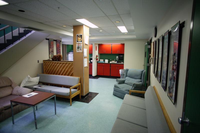 An image of the green room. There are couches and seating and there is a kitchen in the back. To the left of the image there is a staircase.