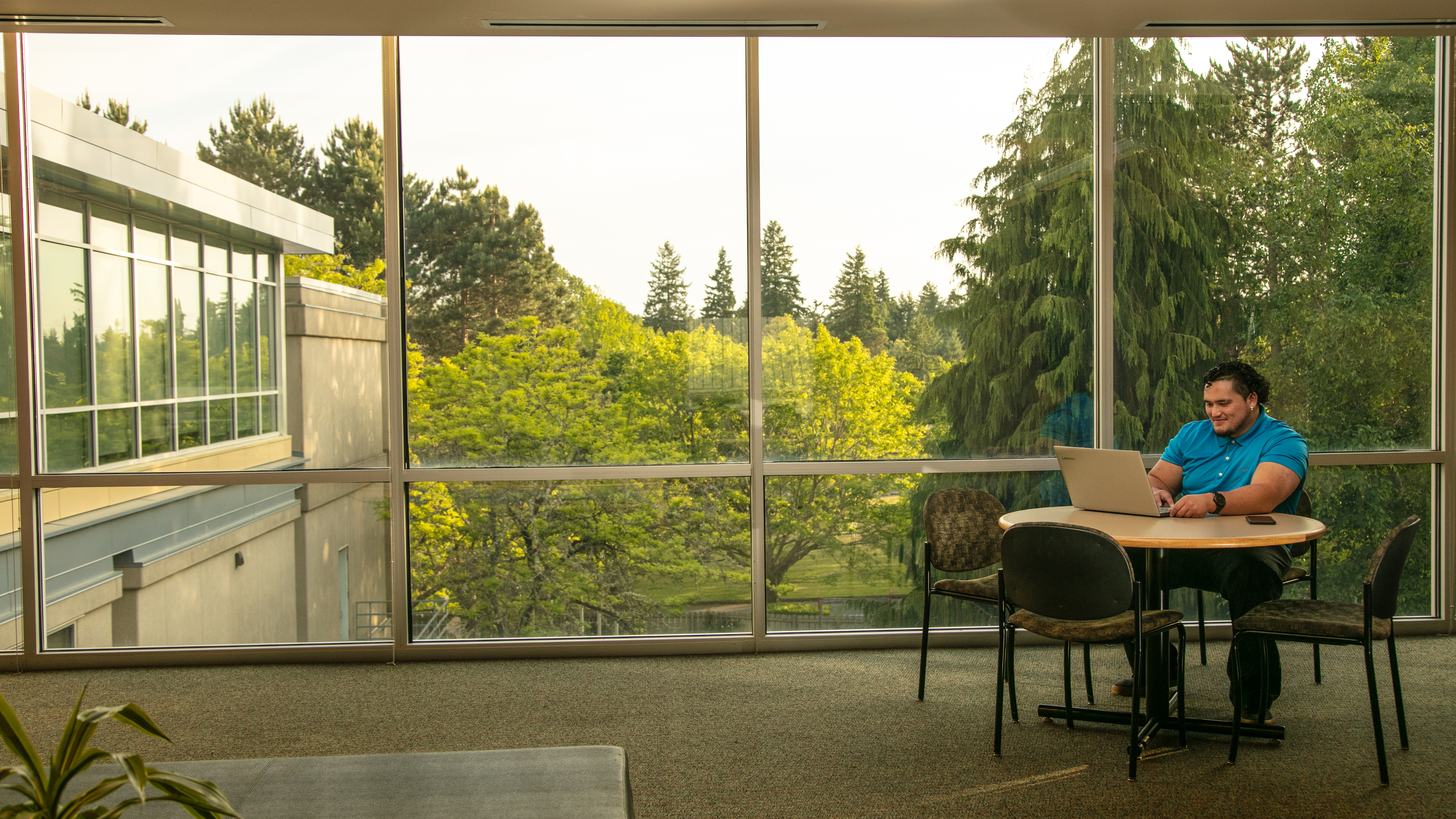 A man sitting at a table in a CWU building. There is a huge window behind him with a view of Evergreen trees. He has his laptop in front of him.