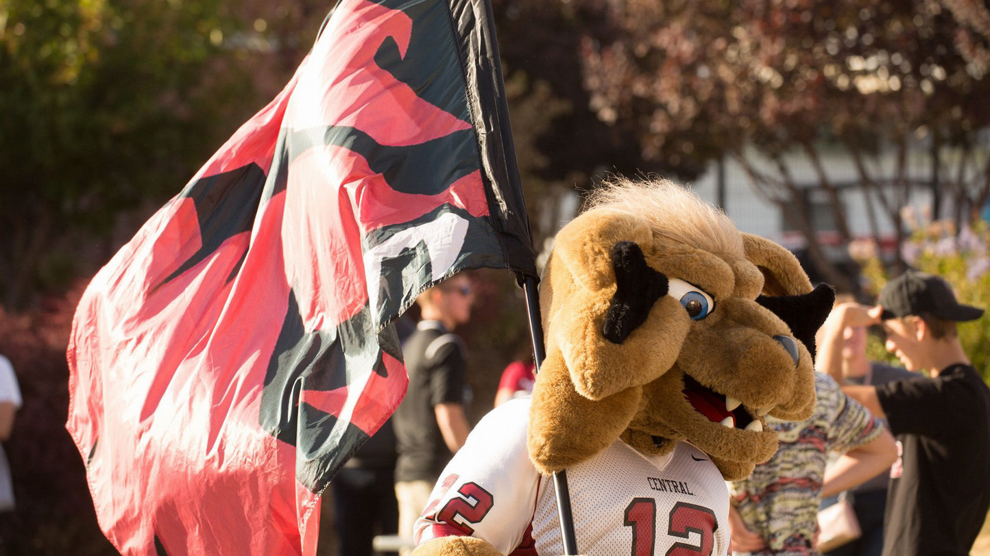 Central Washington University mascot waves the flag. They are outside and there are students in the background