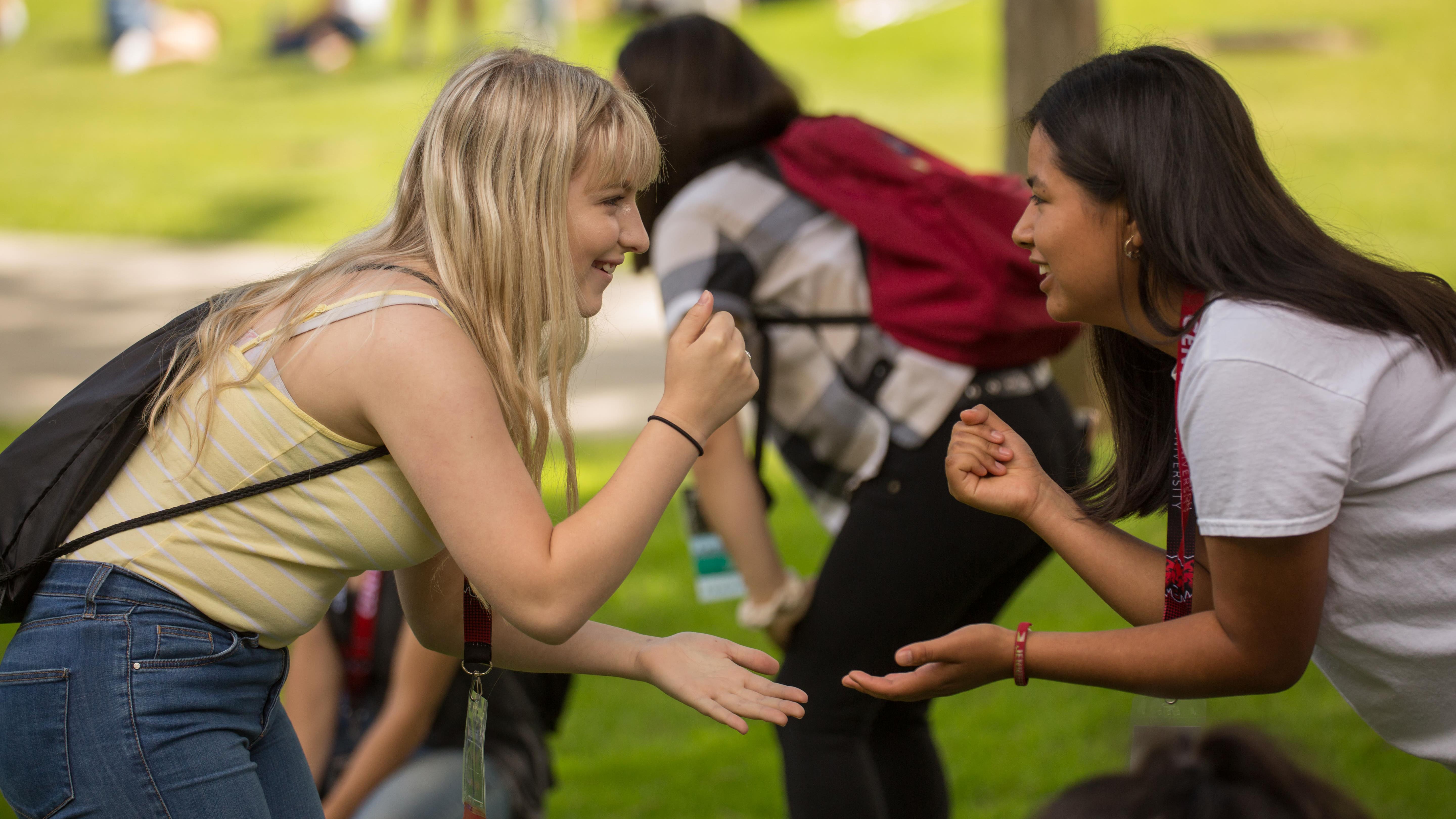 Two students outside playing "rock paper scissors" and they are both holding up a fist for "rock". They are outside and there is green grass in the background.
