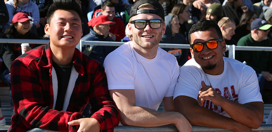 An image of 3 men sitting together on the bleachers. There is a crowd behind them, two of them are wearing sunglasses and white tee shirts and the other man is wearing a red flannel.