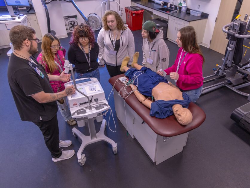 A group of students gathered around a simulated cadaver as a medical professional goes over vital information.