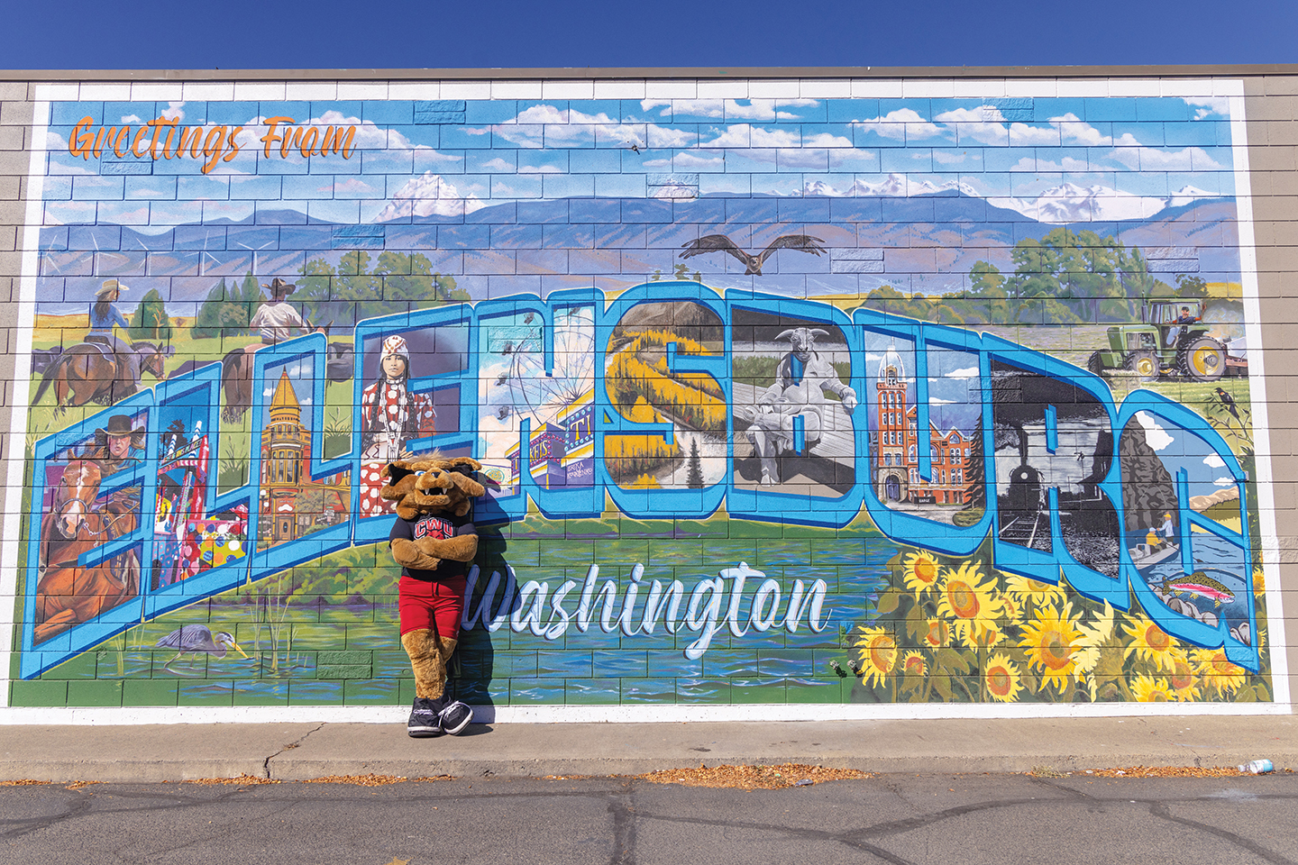 Photo of Wellington the Wildcat standing in front of a downtown Ellensburg mural depicting the town's name.