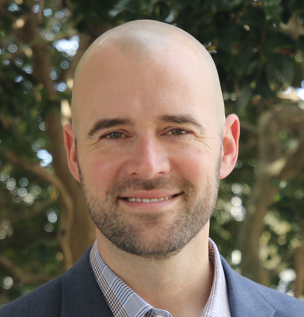 professional headshot of Casey smiling, bald with short cut beard and a grey suit, with a tree in the background