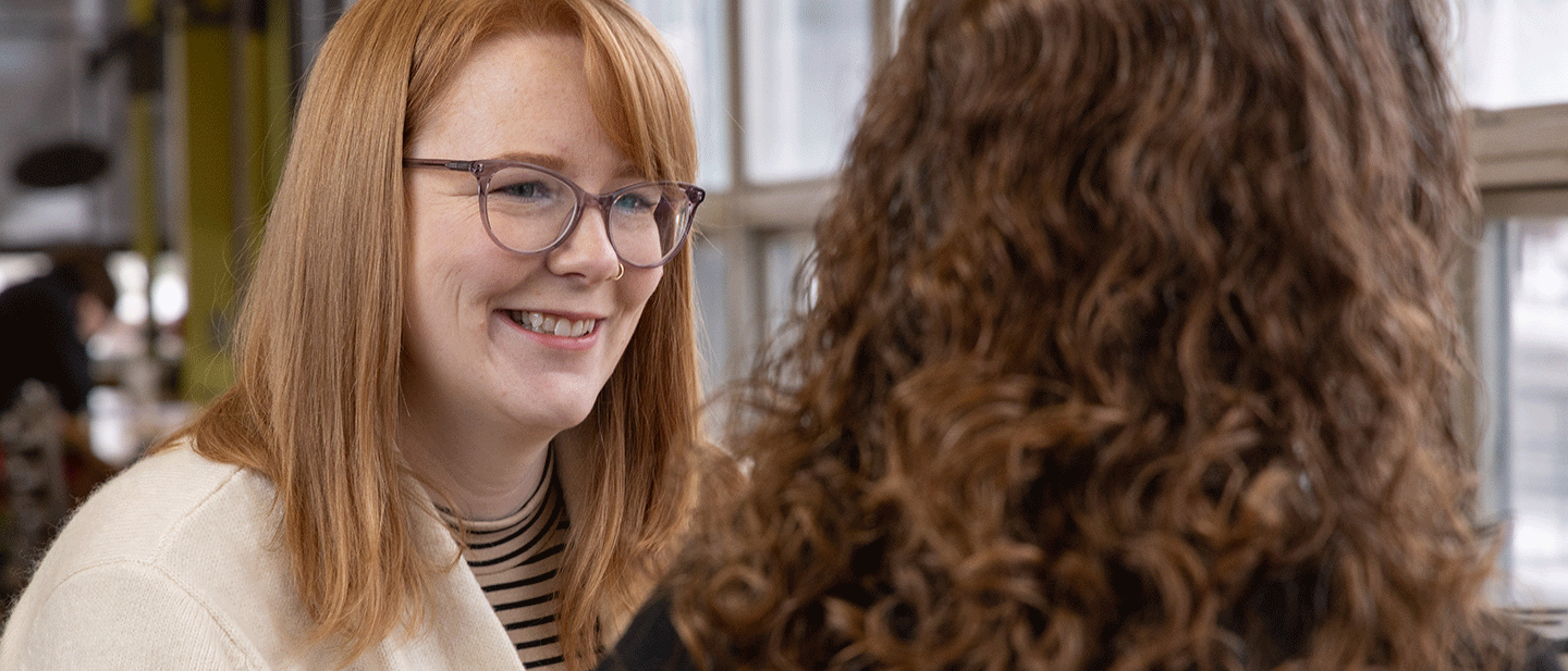 A student with red hair and glasses smiles while talking with another person seated across from them in a well-lit campus space. The image conveys a welcoming, supportive conversation about academic planning and transfer opportunities.