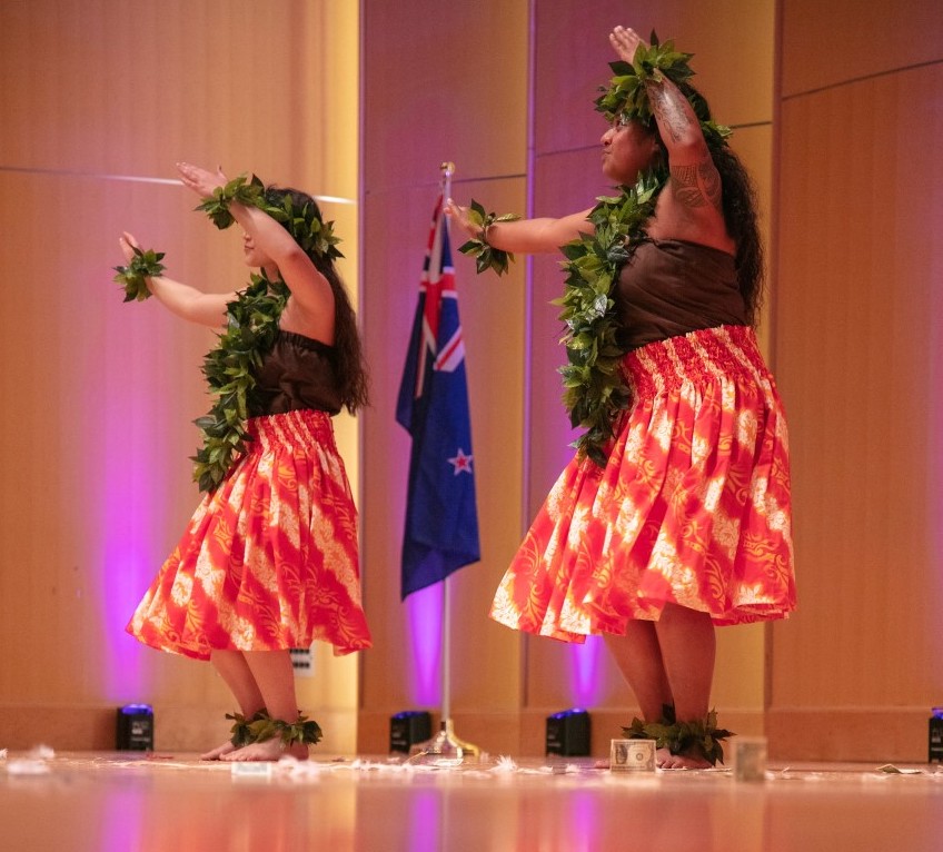 Two individuals on stage wearing long floral skirts with grass wreaths around their neck and wrists preforming a type of dance on stage