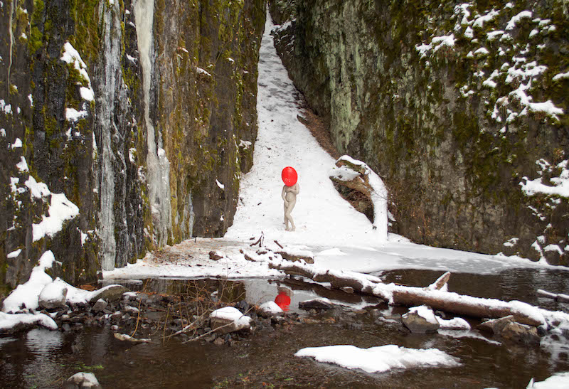 person with an orange ball over their head standing in a snowy nature scene near water.
