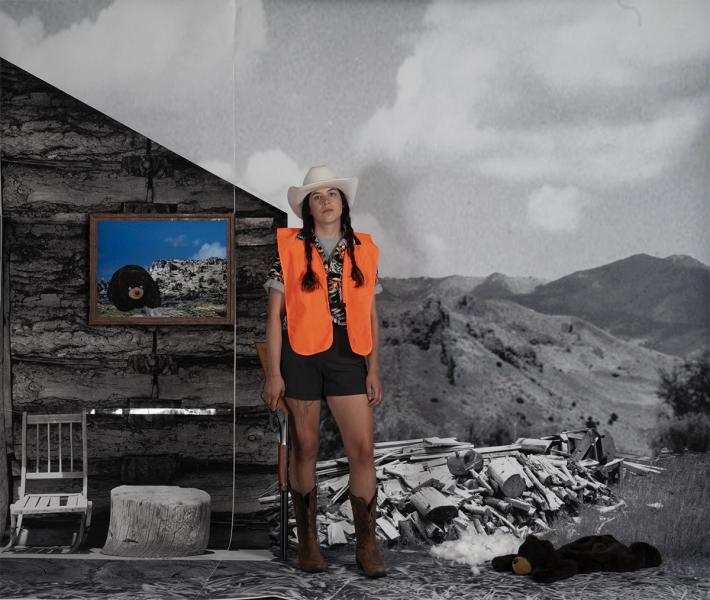 Megan Hansen in a cowboy hat, boots, shorts, and a high visibility orange vest. Standing in a black and white area of an old school cabin with a stuffed bear on the ground.