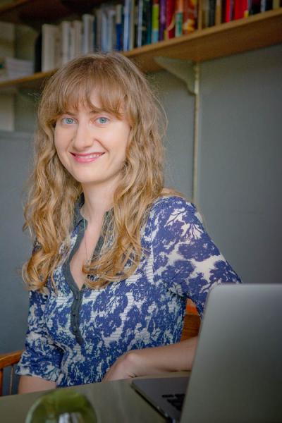woman with blonde curly hair in a blue and white shirt leaning on a desk with a macbook. Woman in the image is Dr. Adair Rounthwaite.