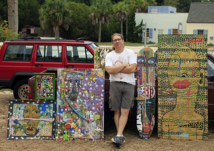 Mike Mitchell stands beside three large paintings that all feature large faces.