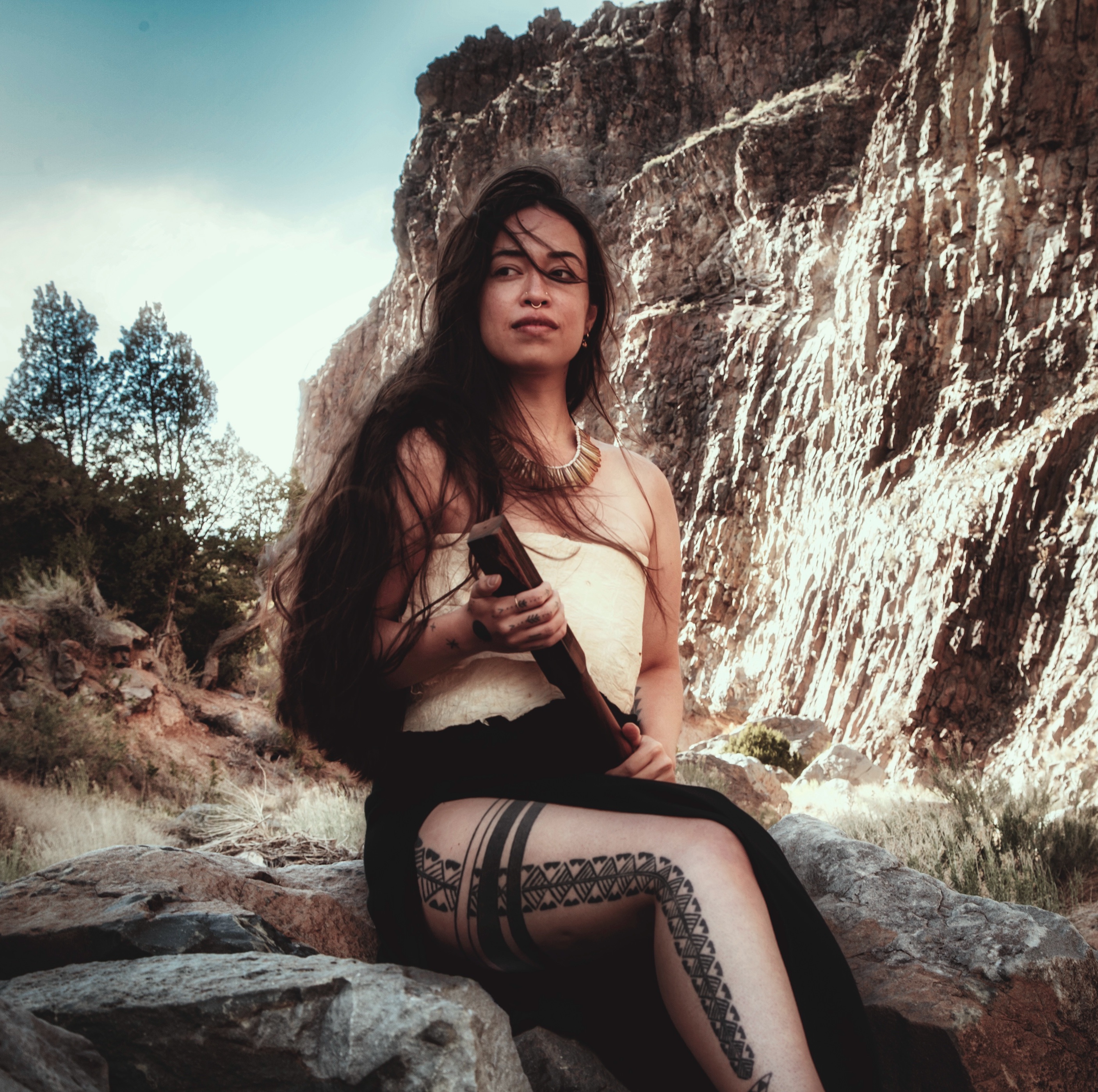 A Hawaiian woman in traditional dress is seated and holding a tool for making bark cloth.
