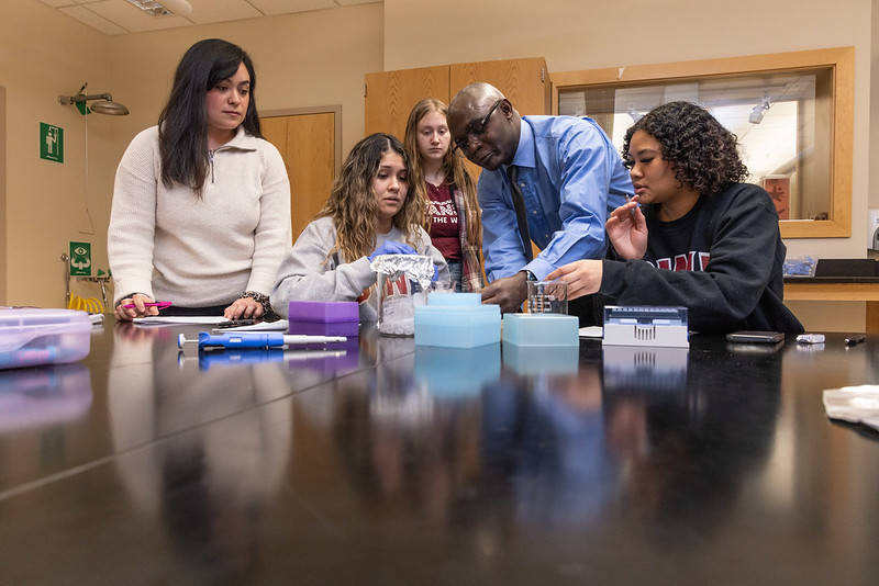 Dr. Dondji instructs four students on proper pipetting techniques as they gather around a dark table with colorful boxes, a pipettor, and glass measuring containers.