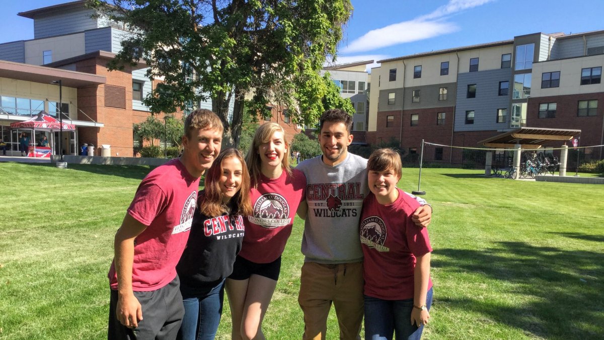 Five CWU students posing with arms wrapped around each other in front of the Barto Hall lawn.