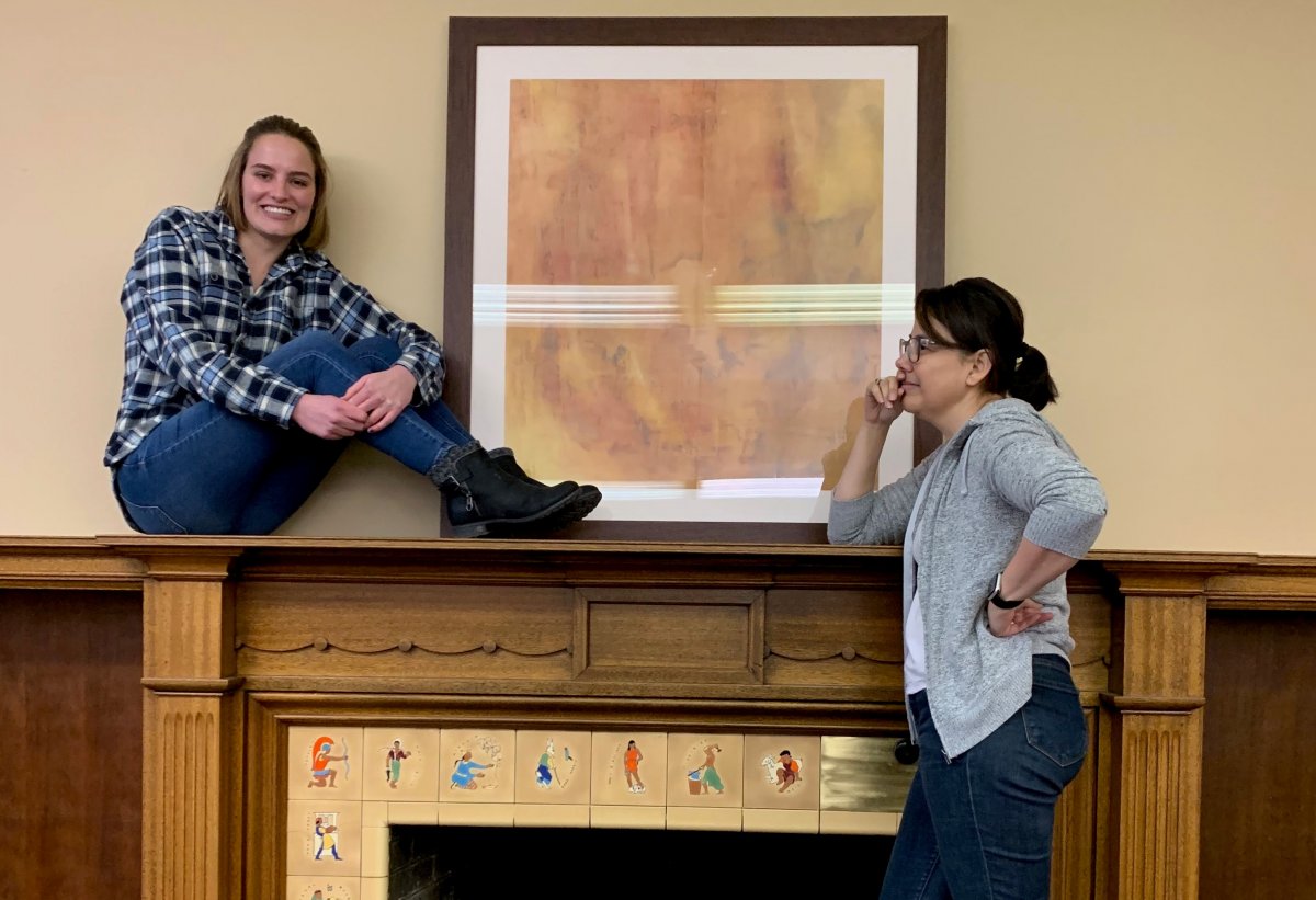 Two Douglas Honors alumni students by a fireplace. One sitting on top, the other resting their elbow on it.
