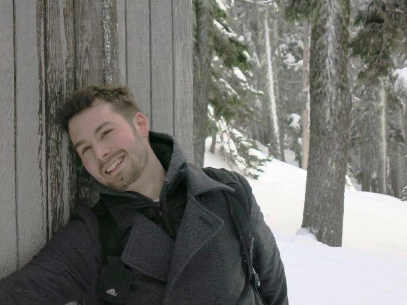 Portrait of Austin Fricke smiling at the camera while leaning against a building with snow and trees in the background