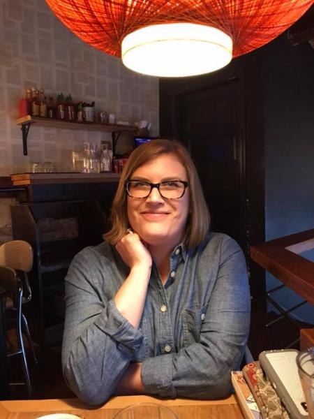 Photo of Seanse Ducken grinning at the camera with one hand up near her face while sitting below a globe lamp