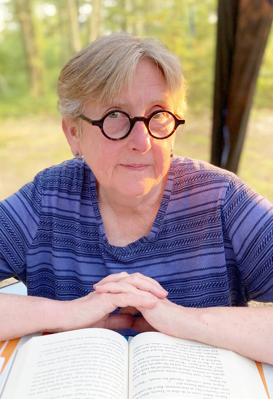 Headshot of Jesse Lee Kercheval sititng with her hands folded in front of her on top of an open book while looking directly at the camera wearing round glasses and a purple striped shirt.