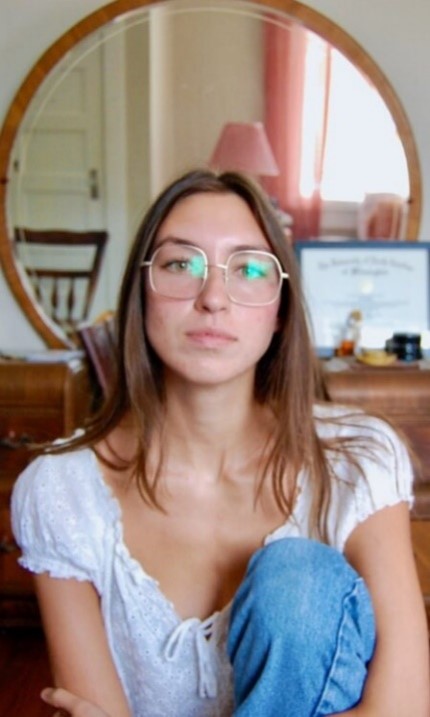 Zoe-Aline Howard facing the camera wearing a white top, blue jeans, and large framed glasses, with a round mirror in the background.