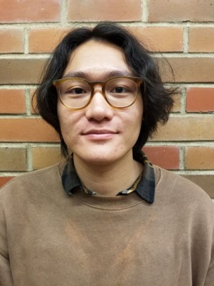 Portrait of Khoa Le in front of a brick wall, wearing glasses and a brown sweater