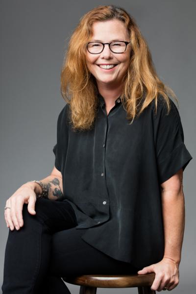 Portrait of Leslie Shipman wearing black glasses and a black outfit sitting on a wooden stool