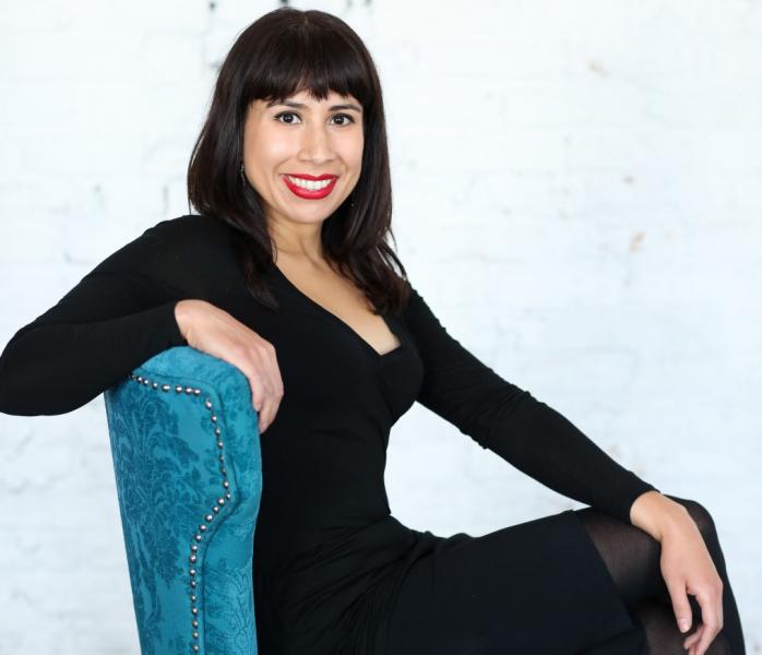 Erika Sanchez smiling, wearing a black dress while sitting on a turquoise chair