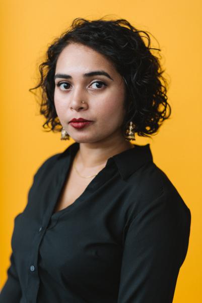 Portrait of SJ Sindu in a black blouse with a yellow background