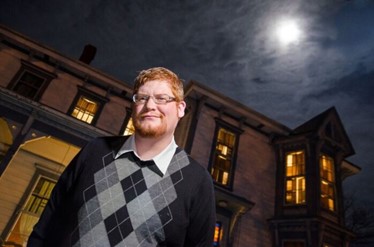 TJ Tranchell is pictured in a black and gray sweater vest in front of a lit up building with the moon shining above him