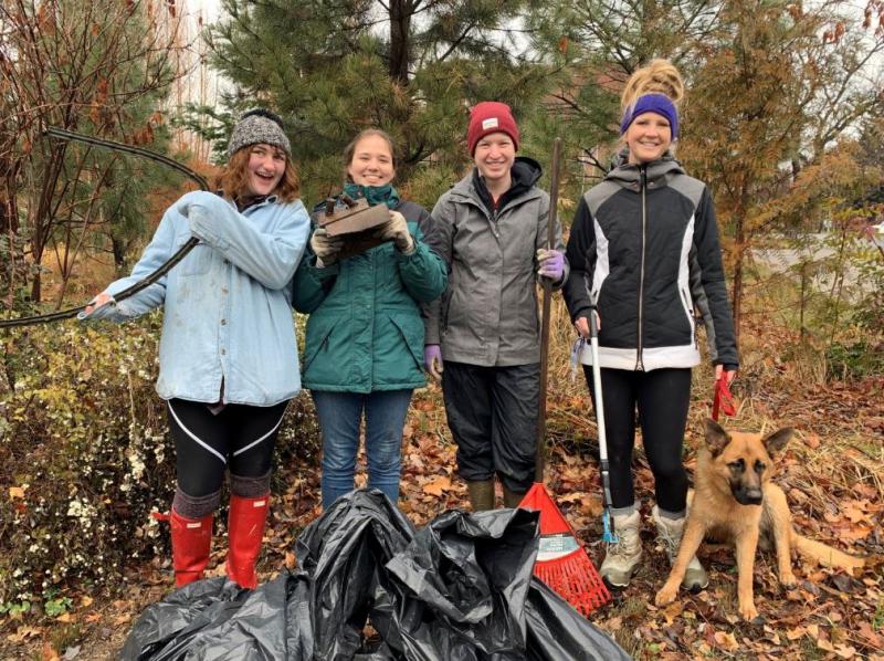 Four members of the CWU Environmental Club wear coats and stand with rakes, black plastic bags, and show some garbage they picked up.  A German Shepard dog lays next to them.