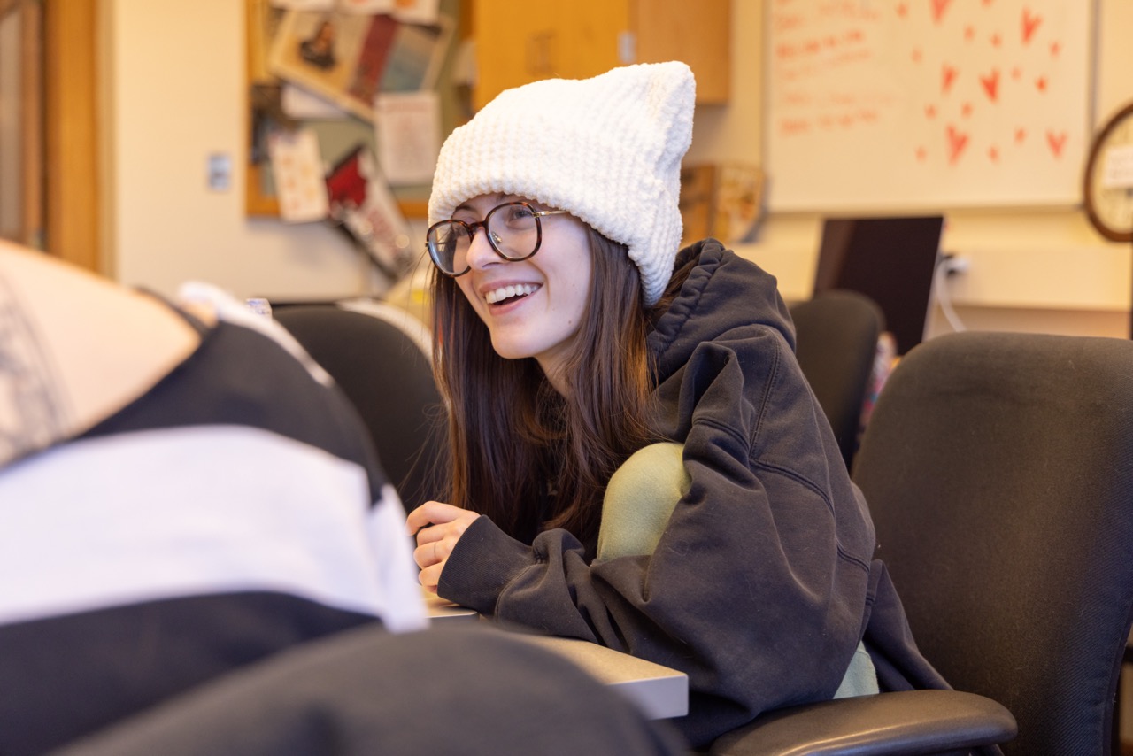 Student in a large beanie and glasses looking at another student during class