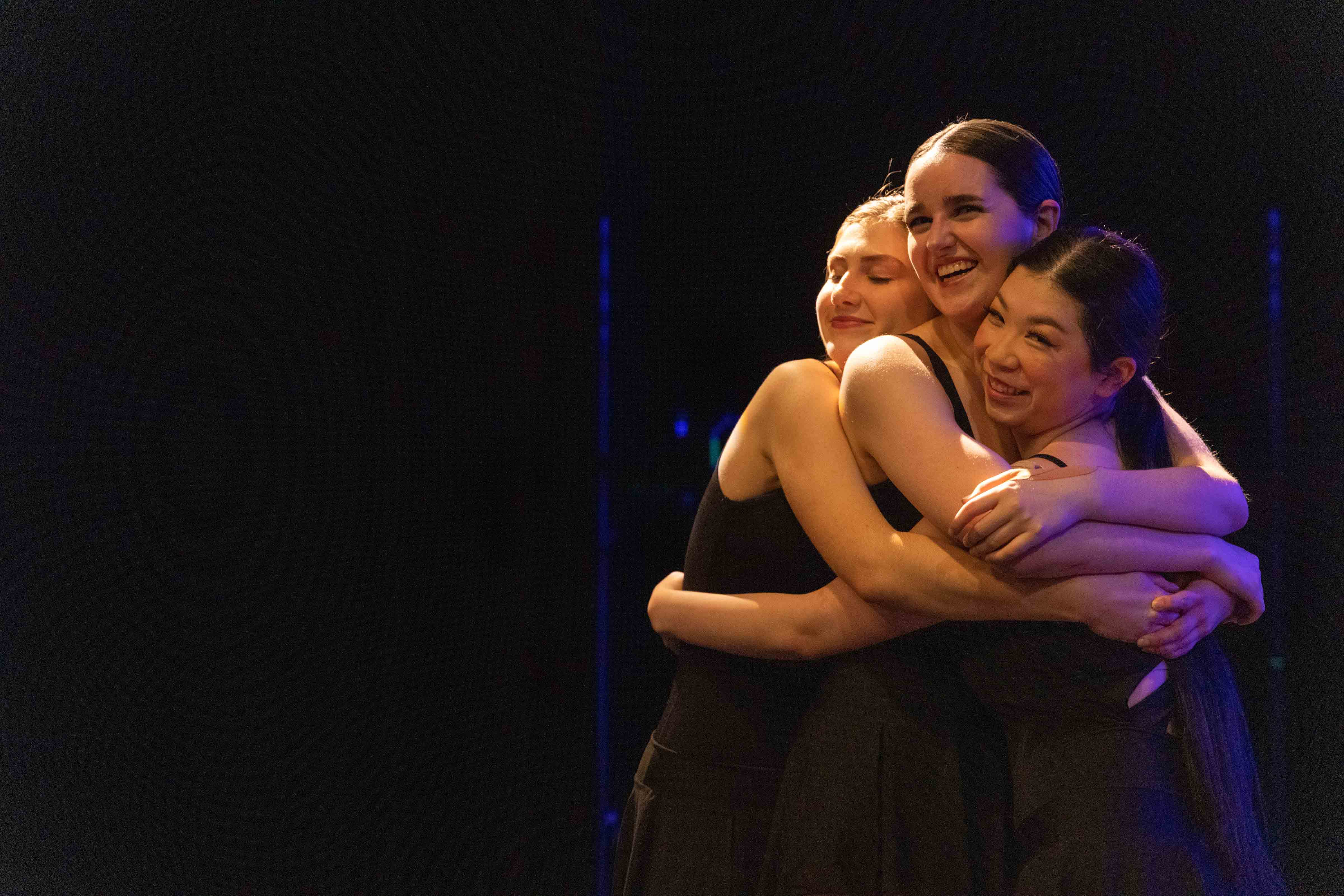 Three dancers smiling and hugging on stage.