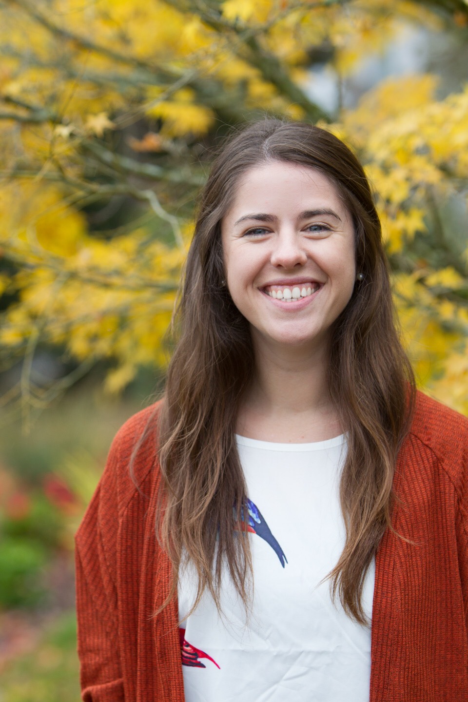 Nina smiling in her headshot. She is wearing a red cardigan and there is a tree behind her