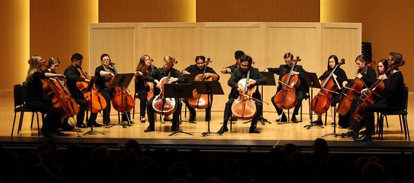Photo of a stage full of cellists in a semi circle with Professor John Michel in the center.