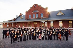 The CWU Symphony Orchestra dressed up in concert black standing in front of the historic Ellensburg train depot at sunset.