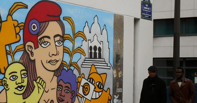 Colorful art on a wall in a city. There are two men walking down the street past the art.