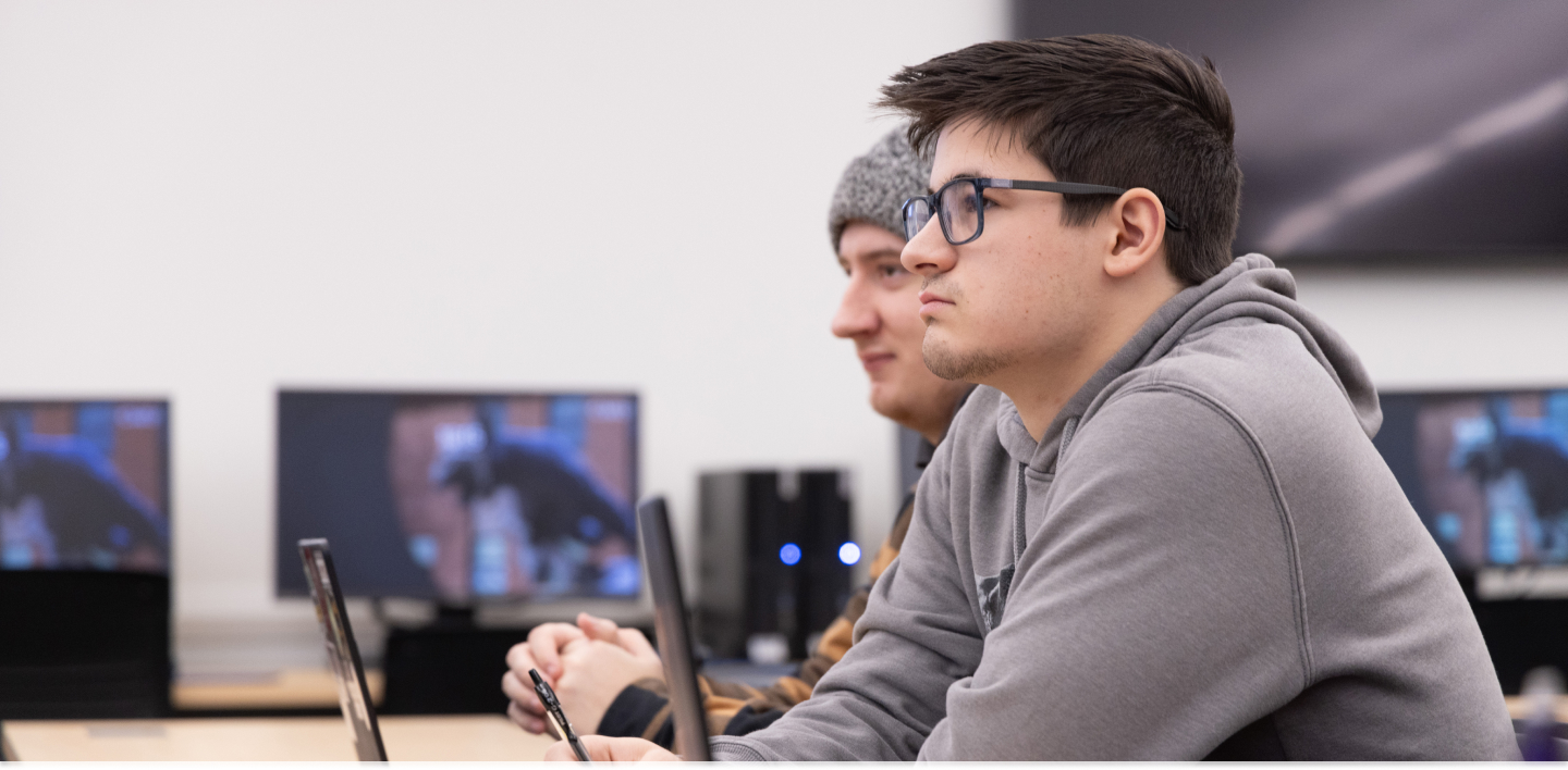 Two students focused on the instructor during class, computer monitors can be seen in the background.