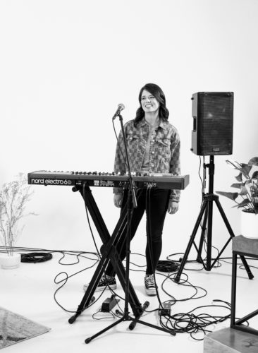 Black and white photo of Ashlyn Nicole Wilson standing in front of an electric keyboard and a microphone, a speaker behind her.