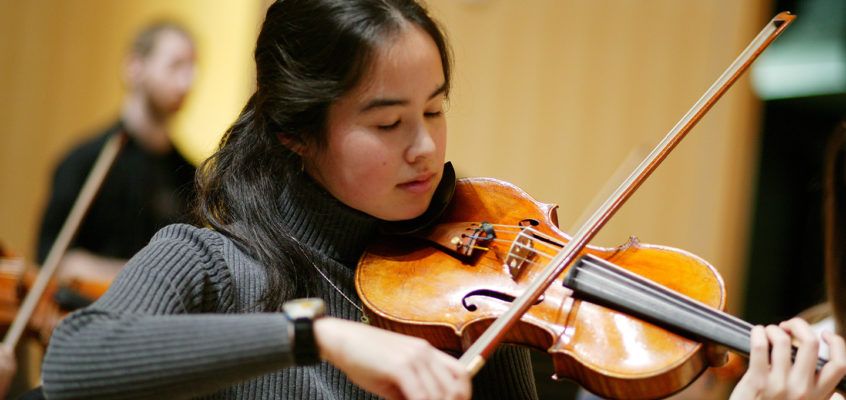 Musician playing the violin in the foreground. Another musician can be seen in the out-of-focus background.