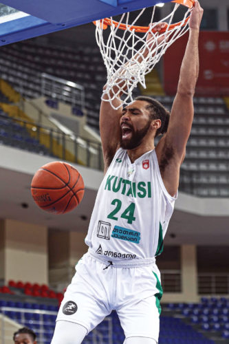 Marqus Gilson hanging off basketball hoop, wearing white uniform, Kutasi 24 written on the front of the jersey