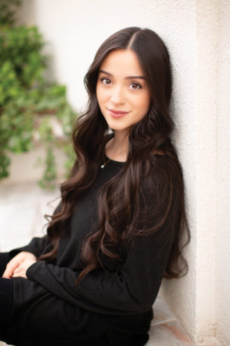 Natalie Velez sitting on the ground and leaning against a wall, looking at the camera smiling.