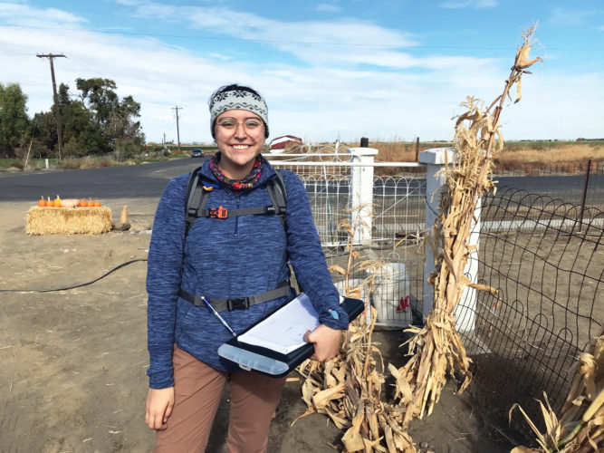 Raelynne Crow standing next to a dried plant, holding a black clipboard with a sheet of paper on top of it.