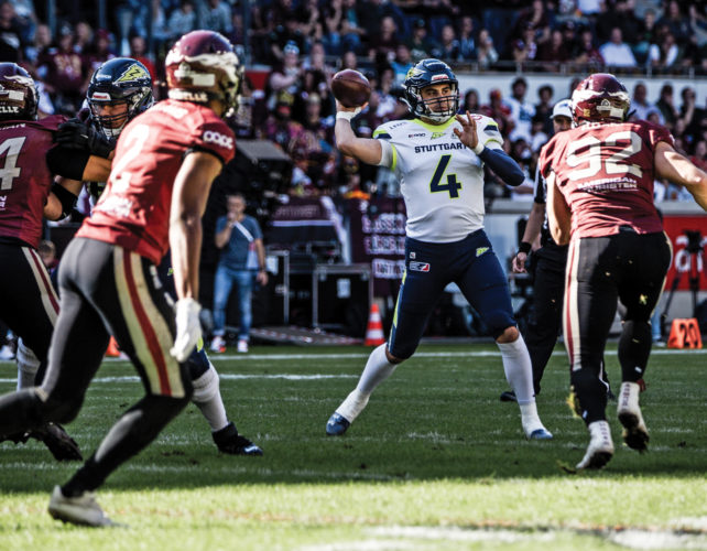 Reilly Hennessey getting ready to throw the football, wearing a white jersey with the number 4 and Stuttgart written on the front, dark blue pants with neon green stripes, other players surround him and one of them is running towards him.