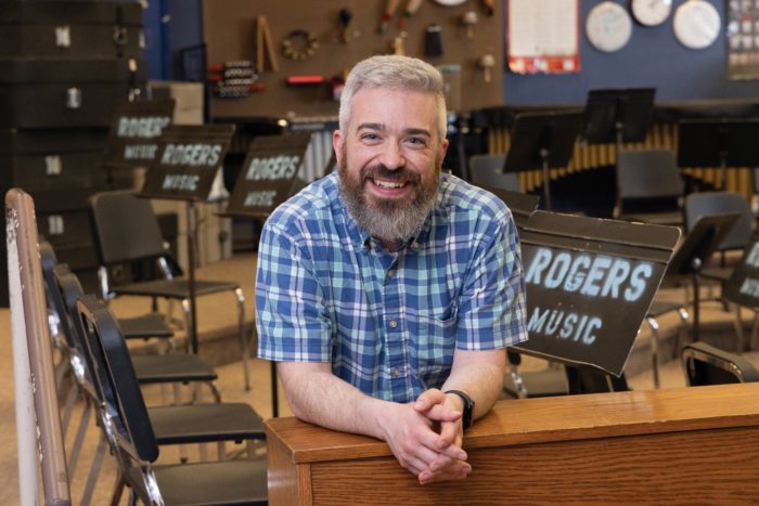 Stephen Pickard leaning against a wooden, short wall, with chairs and music sheet stands behind him.
